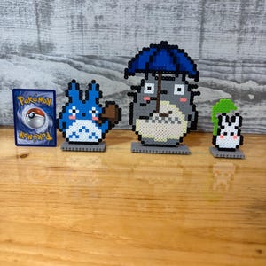 May include: A collection of pixel art figures and a Pok&eacute;mon card. The figures include a blue creature with a brown object, a gray character with a blue umbrella, and a white and green creature. The Pok&eacute;mon card features a red and white Pok&eacute; Ball.