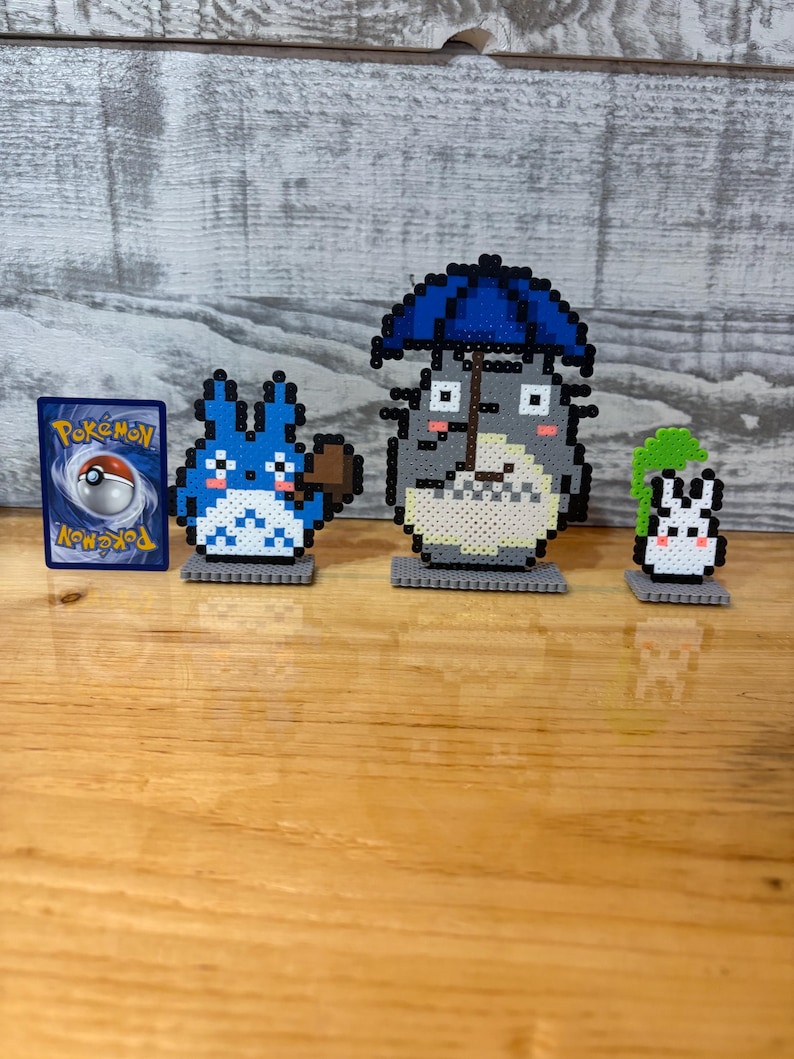 May include: A collection of pixel art figures and a Pok&eacute;mon card. The figures include a blue creature with a brown object, a gray character with a blue umbrella, and a white and green creature. The Pok&eacute;mon card features a red and white Pok&eacute; Ball.