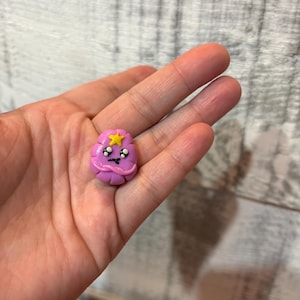 May include: A small, handmade, purple clay charm with a cartoon face. The charm features a yellow star on top and black eyes. The charm is held in a person's hand.