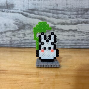 May include: A handmade pixel art zebra figurine with a green leafy top. The zebra is white with black stripes and pink cheeks, standing on a gray base. The figurine is made of small, colorful beads.