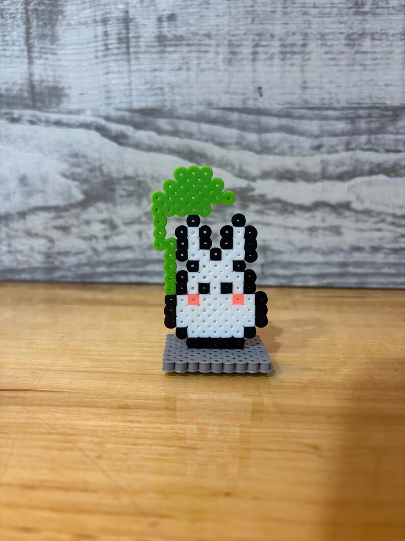 May include: A handmade pixel art zebra figurine with a green leafy top. The zebra is white with black stripes and pink cheeks, standing on a gray base. The figurine is made of small, colorful beads.