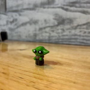Puede incluir: Peque&ntilde;a figura verde de un personaje con ojos grandes y orejas puntiagudas. La figura tiene una capa marr&oacute;n y un cuello beige. La figura est&aacute; sobre una superficie de madera.