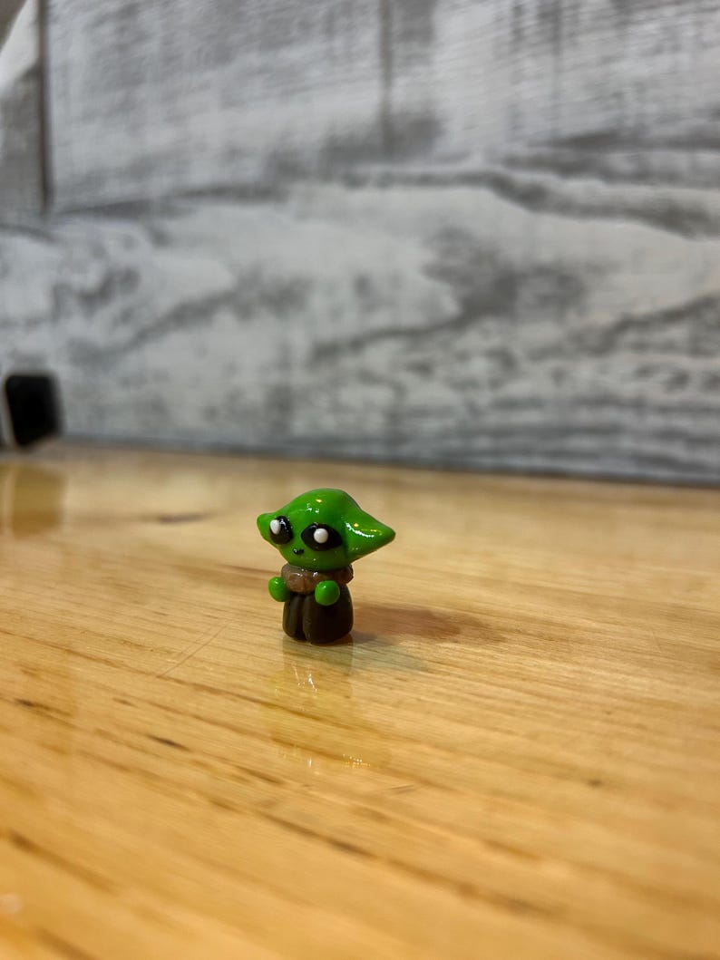 Puede incluir: Peque&ntilde;a figura verde de un personaje con ojos grandes y orejas puntiagudas. La figura tiene una capa marr&oacute;n y un cuello beige. La figura est&aacute; sobre una superficie de madera.