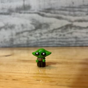 Puede incluir: Peque&ntilde;a figura verde de un personaje con ojos grandes y orejas puntiagudas. La figura tiene una capa marr&oacute;n y est&aacute; sobre una superficie de madera. Es un objeto de colecci&oacute;n.