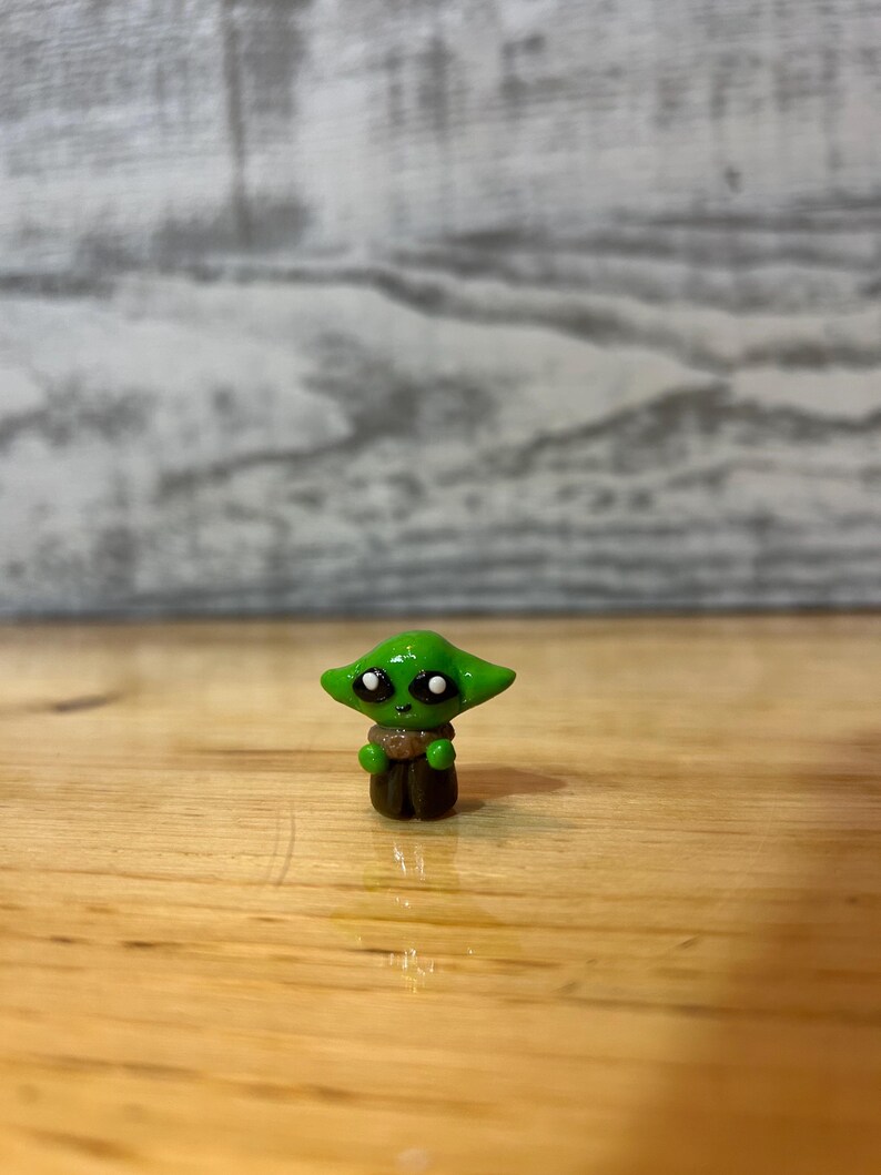 Puede incluir: Peque&ntilde;a figura verde de un personaje con ojos grandes y orejas puntiagudas. La figura tiene una capa marr&oacute;n y est&aacute; sobre una superficie de madera. Es un objeto de colecci&oacute;n.