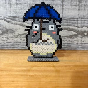 May include: A gray pixel art figure of Totoro holding a blue umbrella. The figure has a white belly, pink cheeks, and black eyes. It is standing on a gray base, and the background is a light gray wood-like surface.