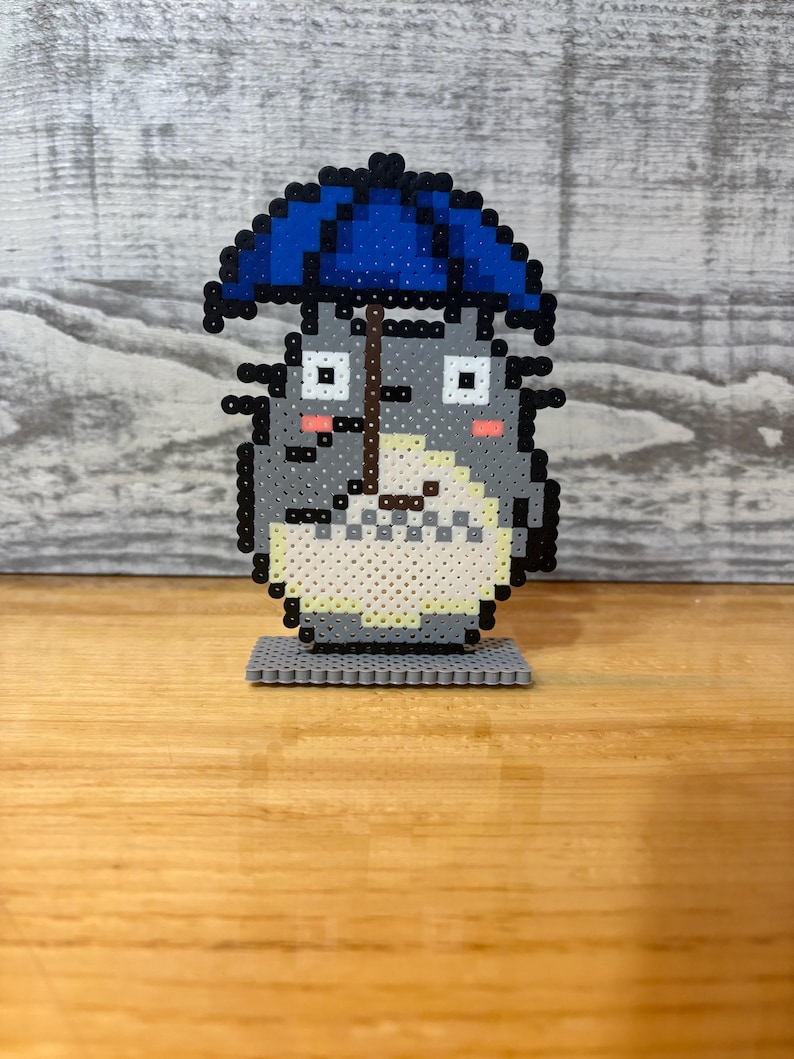 May include: A gray pixel art figure of Totoro holding a blue umbrella. The figure has a white belly, pink cheeks, and black eyes. It is standing on a gray base, and the background is a light gray wood-like surface.