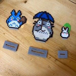 May include: Pixel art figures of characters from the animated film, My Neighbor Totoro. The figures include Totoro with an umbrella, a smaller Totoro, and a Catbus. Also included are three gray rectangular pieces with a slot.
