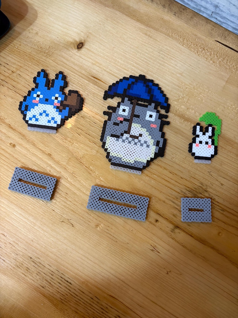 May include: Pixel art figures of characters from the animated film, My Neighbor Totoro. The figures include Totoro with an umbrella, a smaller Totoro, and a Catbus. Also included are three gray rectangular pieces with a slot.