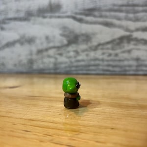 Puede incluir: Peque&ntilde;a figura hecha a mano de una criatura verde con ojos negros y un collar marr&oacute;n. La figura est&aacute; sobre una superficie de madera. La criatura tiene una cabeza redondeada y un cuerpo marr&oacute;n oscuro.