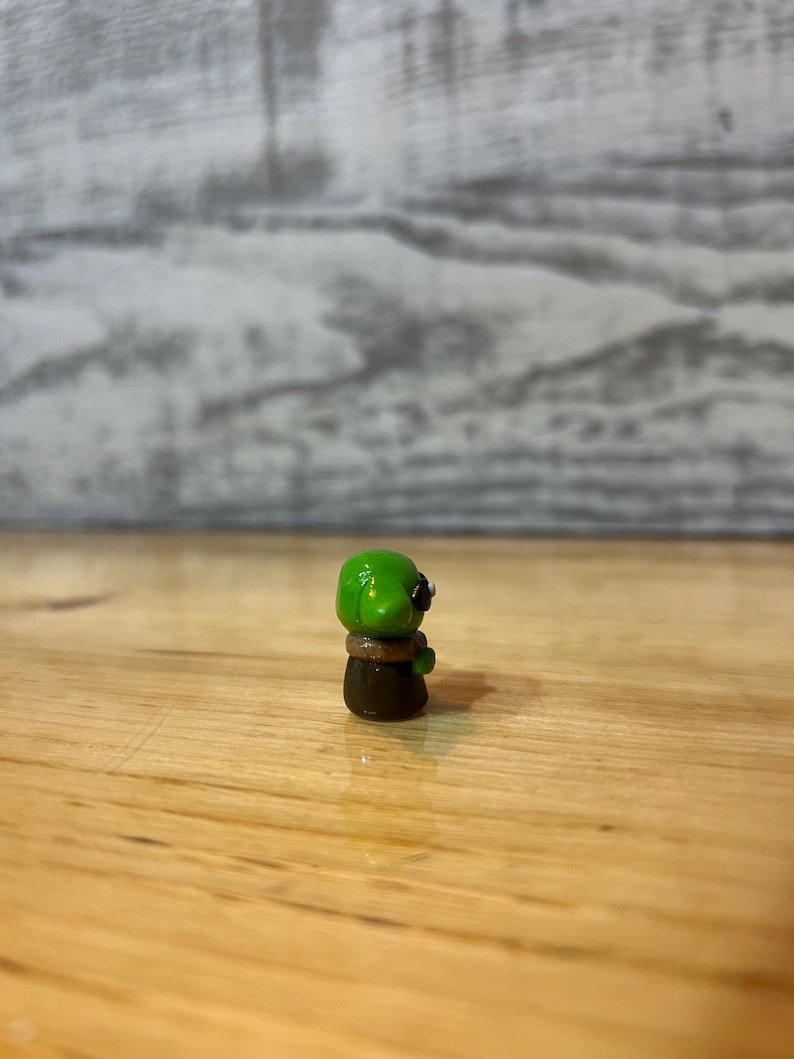 Puede incluir: Peque&ntilde;a figura hecha a mano de una criatura verde con ojos negros y un collar marr&oacute;n. La figura est&aacute; sobre una superficie de madera. La criatura tiene una cabeza redondeada y un cuerpo marr&oacute;n oscuro.