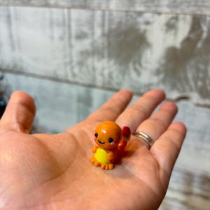 Puede incluir: Una pequeña figura de Charmander naranja con detalles amarillos y una llama roja en la cola. La figura tiene una cara sonriente y se sostiene en una mano abierta. Es un objeto de colección.