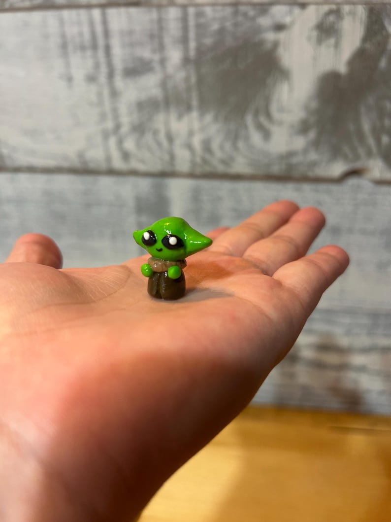 Puede incluir: Peque&ntilde;a figura verde de un personaje con ojos grandes y orejas puntiagudas, que recuerda a una popular franquicia de ciencia ficci&oacute;n. La figura se sostiene en una mano abierta, mostrando su peque&ntilde;o tama&ntilde;o y su detallada artesan&iacute;a. La figura lleva una t&uacute;nica marr&oacute;n.