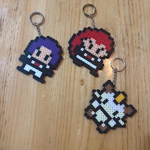 Puede incluir: Se muestran tres llaveros de pixel art. Uno tiene el pelo morado, otro el pelo rojo y el tercero es de diseño marrón y blanco. Cada llavero tiene un llavero plateado adjunto. Los llaveros están hechos de cuentas de plástico de colores.