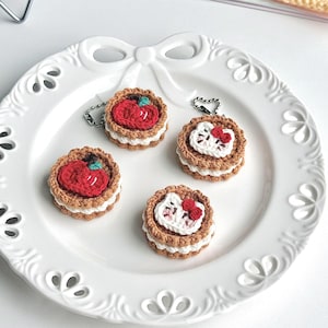 May include: Four crocheted keychains shaped like mini tarts, each with a different design. Two feature red apple designs, and two feature a white cat face with a red bow. The keychains are brown, white, and red, and are displayed on a white decorative plate.