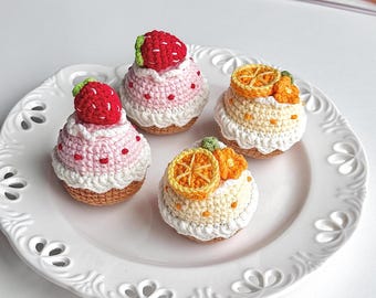 May include: Four crocheted cupcakes on a white plate. Two cupcakes are topped with red strawberries, and two are topped with orange slices. The cupcakes have white frosting and brown bases. The plate has a decorative edge.