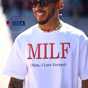 Può includere: Maglietta bianca con la scritta "MILF" a blocchi rossi e la frase "(Man, I Love Ferrari)" sotto. La maglietta è indossata da una persona con occhiali da sole e una collana di perline colorate. È visibile un logo "Best Quality USA".
