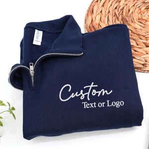 Può includere: Una felpa blu navy con colletto con zip a un quarto. La parola "Custom" è ricamata in corsivo bianco, con "Text or Logo" sotto. La felpa è piegata, con un'etichetta visibile.