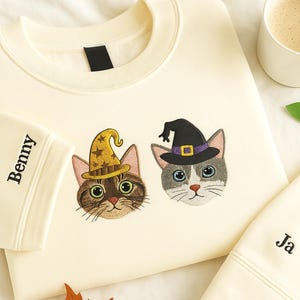 以下が含まれることがあります： クリーム色のスウェットシャツ。魔女の帽子をかぶった猫の顔が刺繍されています。片方の猫は金の星模様の帽子、もう片方は紫色のバンドが付いた黒い帽子をかぶっています。袖には黒い文字で「Benny」と書かれています。