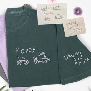 May include: A selection of t-shirts in green, lilac, and grey. The front shirt is a dark green colour, featuring white embroidered text and drawings. The text says "Poppy" with a bicycle and train. A sticky note reads "Left Chest Poppy".