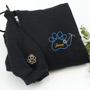 May include: Black sweatshirt with a zipper and drawstring. The sweatshirt features a paw print design with the name "James" embroidered in gold and blue. A stethoscope is also embroidered on the paw print.