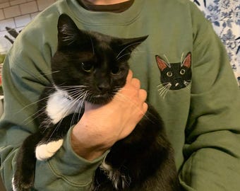 Puede incluir: Una sudadera de cuello redondo color verde oliva con un gráfico bordado de una cara de gato negro en el pecho izquierdo. El diseño del gato incluye bigotes blancos, orejas rosadas por dentro y ojos grandes. Tiene mangas largas.