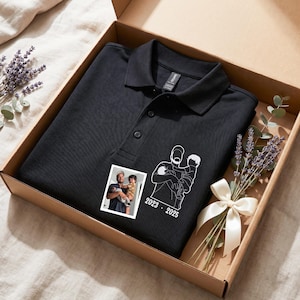 Könnte beinhalten: Ein schwarzes Poloshirt mit einer weißen Grafik einer Person, die ein Kind hält, und den Jahren 2023-2025. Das Shirt liegt in einem Karton, mit einem kleinen Foto und Lavendelzweigen. Ein weißes Band schmückt den Lavendel.