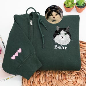 May include: Dark green zip-up hoodie with an embroidered cat face and the name "Bear" in white. The sleeve features three pink hearts. A circular image of a cat is in the upper right corner.