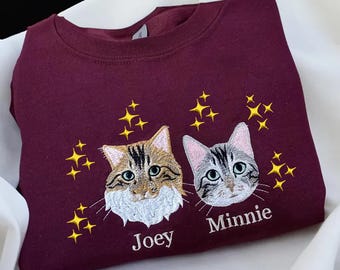 Puede incluir: Una sudadera de cuello redondo color granate con retratos bordados de dos gatos. El gato de la izquierda es marrón y blanco, etiquetado como Joey, y el de la derecha es gris y blanco, etiquetado como Minnie. Estrellas amarillas rodean las cabezas.