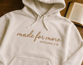 May include: Cream-colored hoodie with a front pocket and drawstring hood. The phrase "made for more" and the reference "Ephesians 2:10" are embroidered in a brown script across the chest. A Bible is open in the background.