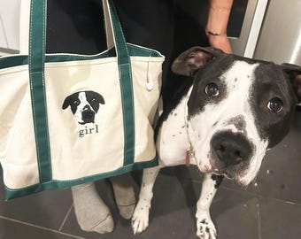 May include: A cream-colored canvas tote bag with dark green trim and handles. The front of the bag features an embroidered black and white dog face graphic above the lowercase text "girl" in a simple black serif font, centered on the bag.