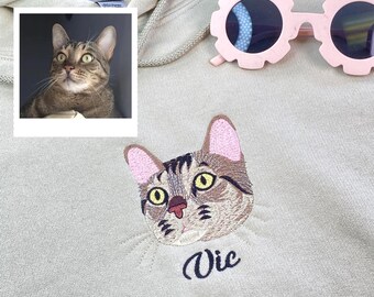 Puede incluir: Una sudadera con capucha beige claro con un gato marrón y negro bordado en la parte delantera. El nombre del gato, "Vic", está bordado debajo de la imagen del gato.