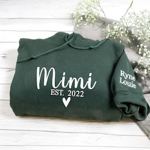 May include: A dark green hoodie with the word "Mimi" in white script, "EST. 2022" below, and a white heart. The sleeve cuff has the text "Ryne Louie". The hoodie is folded on a light wood surface.