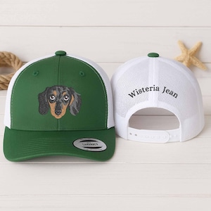 May include: A green and white trucker hat with an embroidered dachshund dog. The hat has a green peak and crown with a white mesh back. The text "Wisteria Jean" is embroidered on the back of the hat.