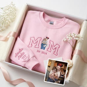 Könnte beinhalten: Ein rosa Sweatshirt und ein passender Ärmel mit gesticktem Text und einer Grafik einer Mutter und eines Kindes. Auf dem Sweatshirt steht "MOM Est 2024". Der Ärmel ist mit einem Namen und einem Herzsymbol personalisiert. Die Artikel befinden sich in einer Geschenkbox.