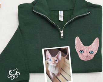 Puede incluir: Una sudadera verde oscuro con cremallera y una cara de gato bordada en rosa en la parte delantera. La sudadera tiene una huella de pata bordada en blanco en la manga izquierda. Una foto de un gato sin pelo es visible en el fondo.