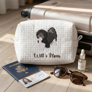 May include: A white waffle-textured fabric pouch featuring an embroidered black and white dog and the text Will's Mom in brown script. The pouch has side tabs and is positioned near a passport, an identification card, sunglasses, and a serum bottle.