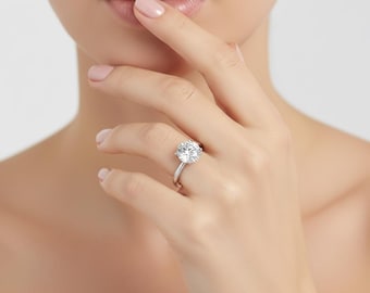Könnte beinhalten: Ein silberner Verlobungsring mit einem großen, runden, klaren Edelstein. Der Ring ist auf einem schmalen Band gefasst und wird an einem Finger gezeigt. Der Hintergrund ist eine weiche, neutrale Farbe.