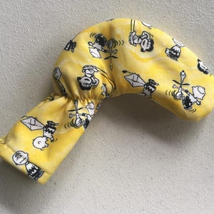 May include: A yellow golf club head cover featuring a repeating pattern of black and white Peanuts comic strip characters including Charlie Brown, Lucy, and Snoopy. The fabric cover has a curved shape with an elastic opening at the base.