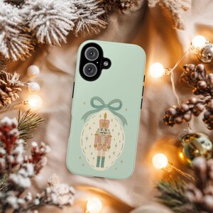Puede incluir: Funda de teléfono verde menta con un diseño festivo de cascanueces. La ilustración está dentro de un marco ovalado, adornado con un lazo y estrellas. La funda tiene una configuración de doble cámara y detalles negros, ideal para regalos navideños.