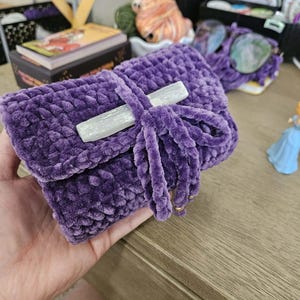 May include: A deep purple crochet pouch made from soft, plush yarn. The rectangular pouch features a fold-over flap secured by a matching purple crocheted tie, which is accented with a long, translucent white selenite crystal wand.