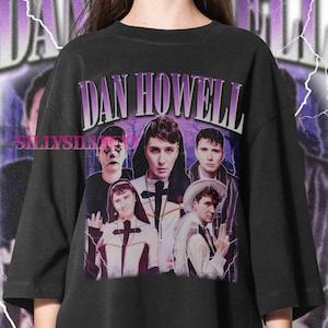 Puede incluir: Camiseta negra con un diseño gráfico de estilo vintage de Dan Howell en morado y blanco. El texto "DAN HOWELL" se muestra de forma destacada. La camiseta tiene cuello redondo y mangas cortas.