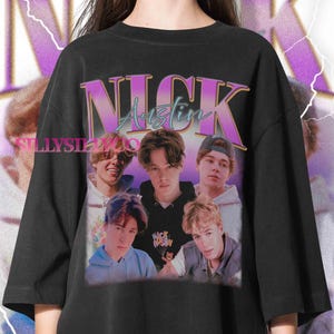 May include: Black crew neck t-shirt featuring a graphic of five young men with the text "NICK Austin" in a retro style. The graphic has a purple and teal color scheme. The shirt is a casual style.
