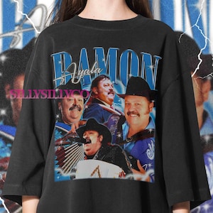May include: Black t-shirt featuring a collage of images of a man with a mustache and a cowboy hat. The name "Damon Ayala" is displayed in blue text above the images. The shirt has a vintage, retro style.