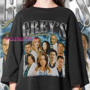 May include: Black t-shirt featuring a graphic of the Grey's Anatomy cast with the show's logo in silver lettering. The design includes a collage of character portraits against a blue and white backdrop.