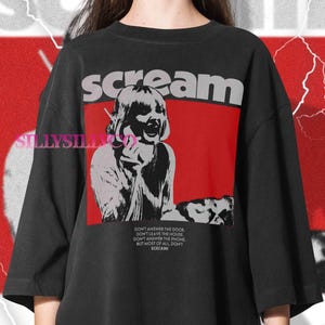 May include: Black graphic t-shirt with the word "scream" in gray letters above a red rectangle. The graphic features a distressed image of a person screaming into a phone. Text below reads "Don't answer the door..." and "Scream!"