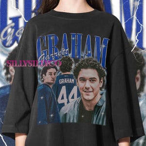 May include: A black t-shirt featuring a graphic print with the text "GRAHAM" and "Garrett" in blue. The design includes two images of a person, one showing the back of a jersey with the name "GRAHAM" and the number "44", accented by star icons.