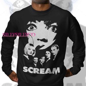 May include: Black sweatshirt with a white graphic from the film "Scream." The design features a large face with wide eyes, a knife, and the word "SCREAM." Below the face are images of the movie's cast. A classic horror movie design.