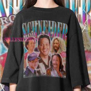 May include: Black t-shirt featuring a collage of images of Rob Schneider with the name "SCHNEIDER" in a retro font. The shirt has a vintage aesthetic, with a distressed look and a colorful design. The text "ROB" is also included.