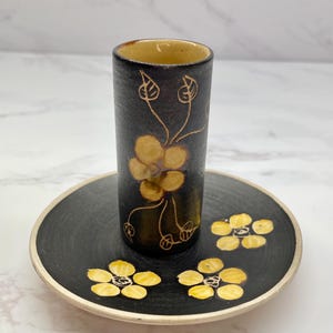 May include: A black ceramic shot glass and saucer set. The shot glass and saucer are decorated with yellow flowers and brown stems. The rim of the saucer is cream-coloured.
